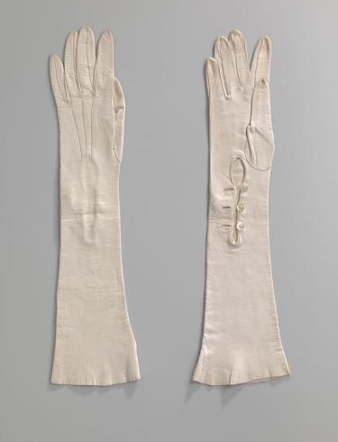 Paar lange dameshandschoenen van wit glacéleer. Op de hand drie stiksels. Bij de pols sluitend middels drie witte kunststof knopen. Amsterdam Museum (objectnummer KA 17210). 