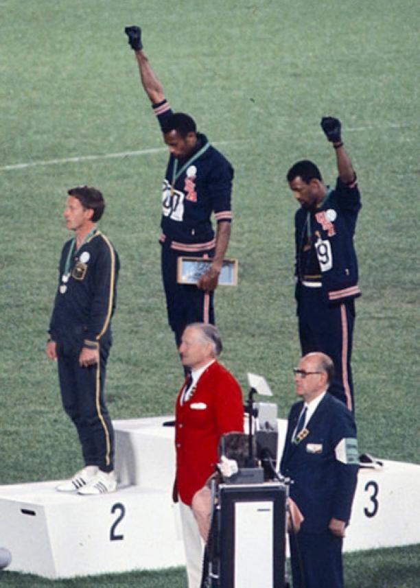 Medaillewinnaars Tommie Smith (goud), Peter Norman (zilver) en John Carlos (brons) staan op het podium van de Olympische Spelen, 1968. Smith en Carlos houden een gebalde vuist in de lucht als mensenrechten- of Black Power-saluut. Bron: Wikipedia.